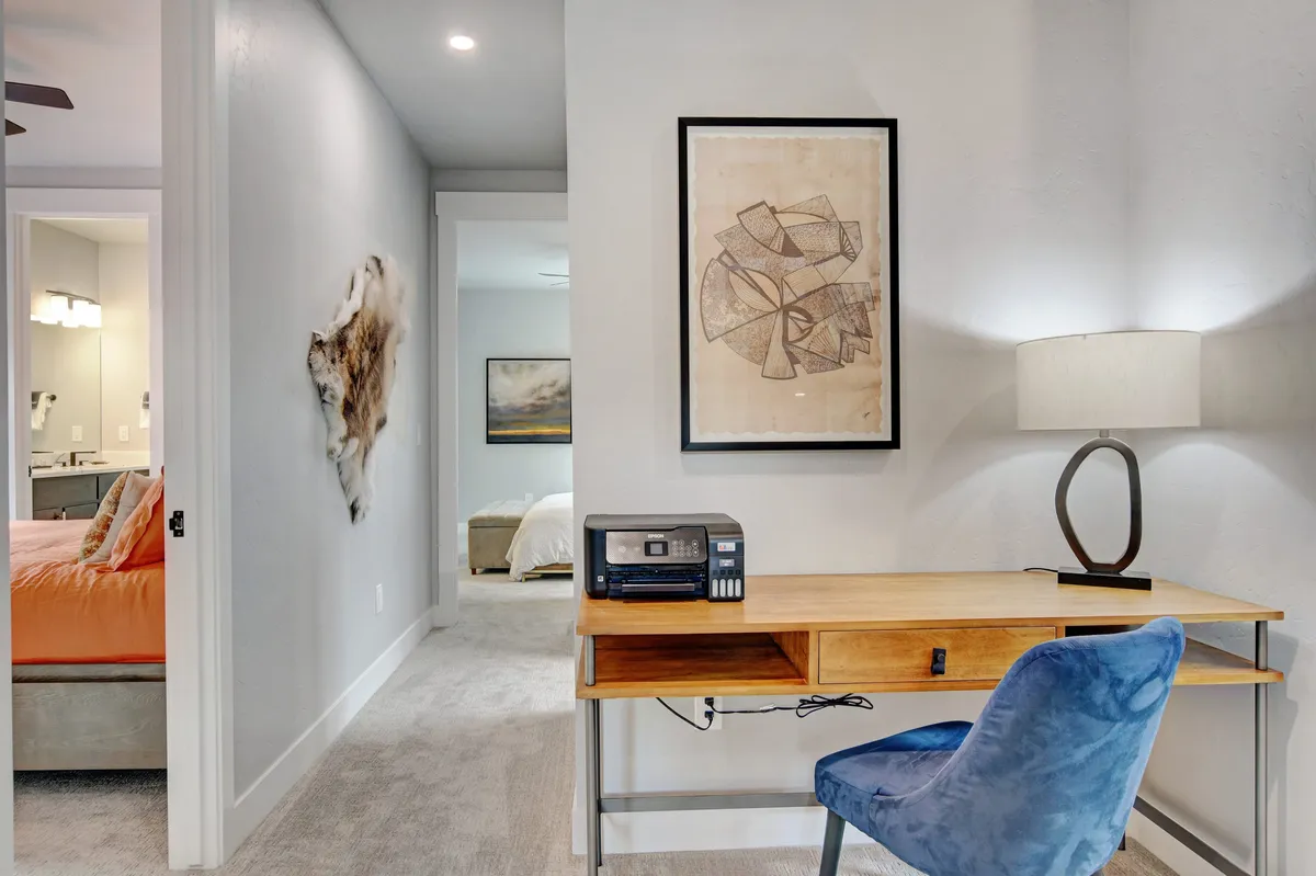 A cozy, dedicated workspace with a wooden desk, comfortable blue chair, framed artwork, and a lamp, set in a quiet corner that still feels connected to the main living areas.