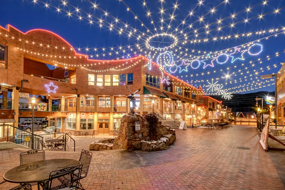 A festive exterior scene with string lights, decorative stars, a brick walkway, outdoor seating, and a fountain. People are seen dining, shopping, and enjoying a lively atmosphere, suggesting an active, inviting mountain town vibe.