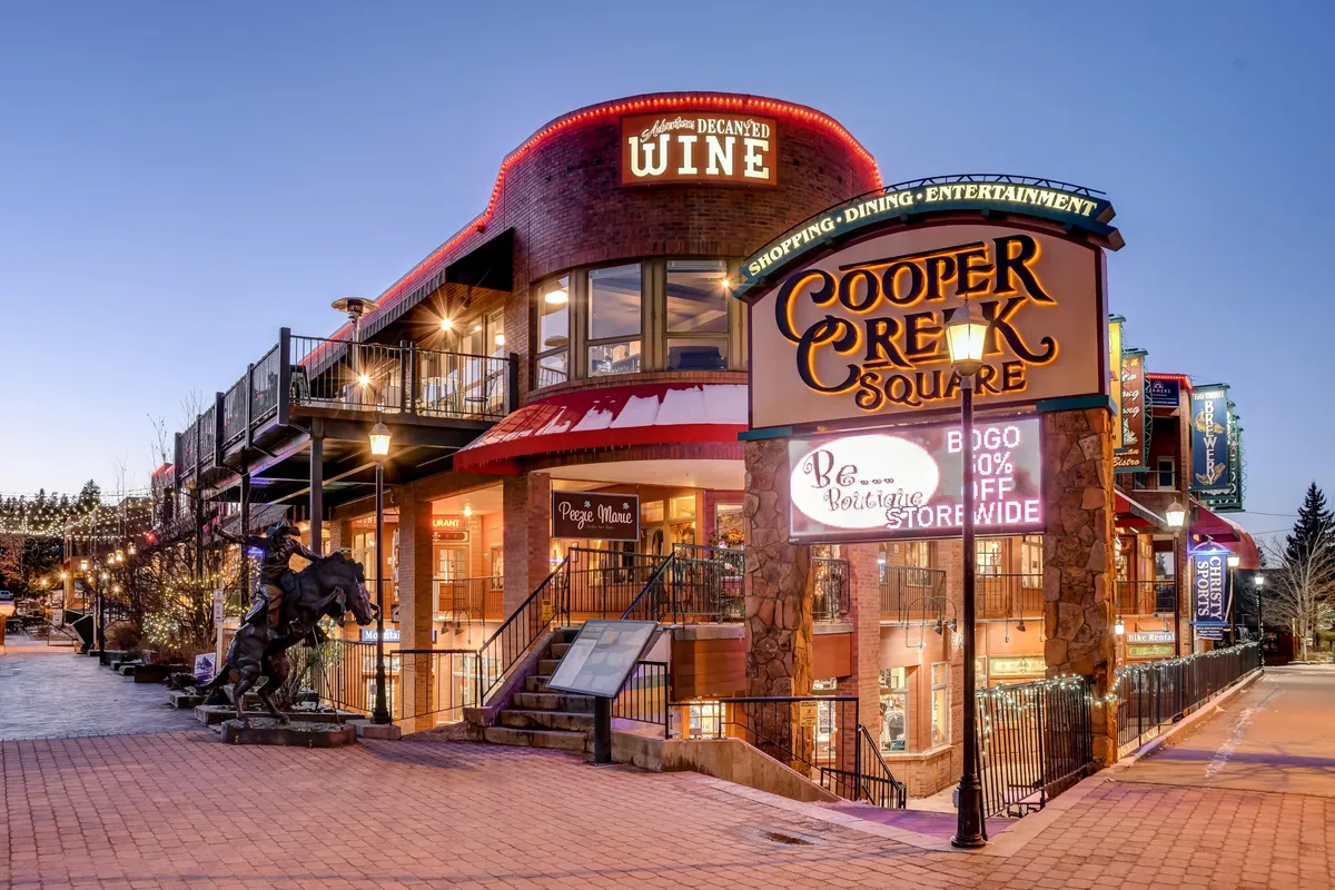The image features Cooper Creek Square with its prominent sign, a wine shop sign, inviting outdoor dining spaces, and a decorative statue. The area is bustling with activity, embodying the heart of downtown Winter Park's social and commercial scene.