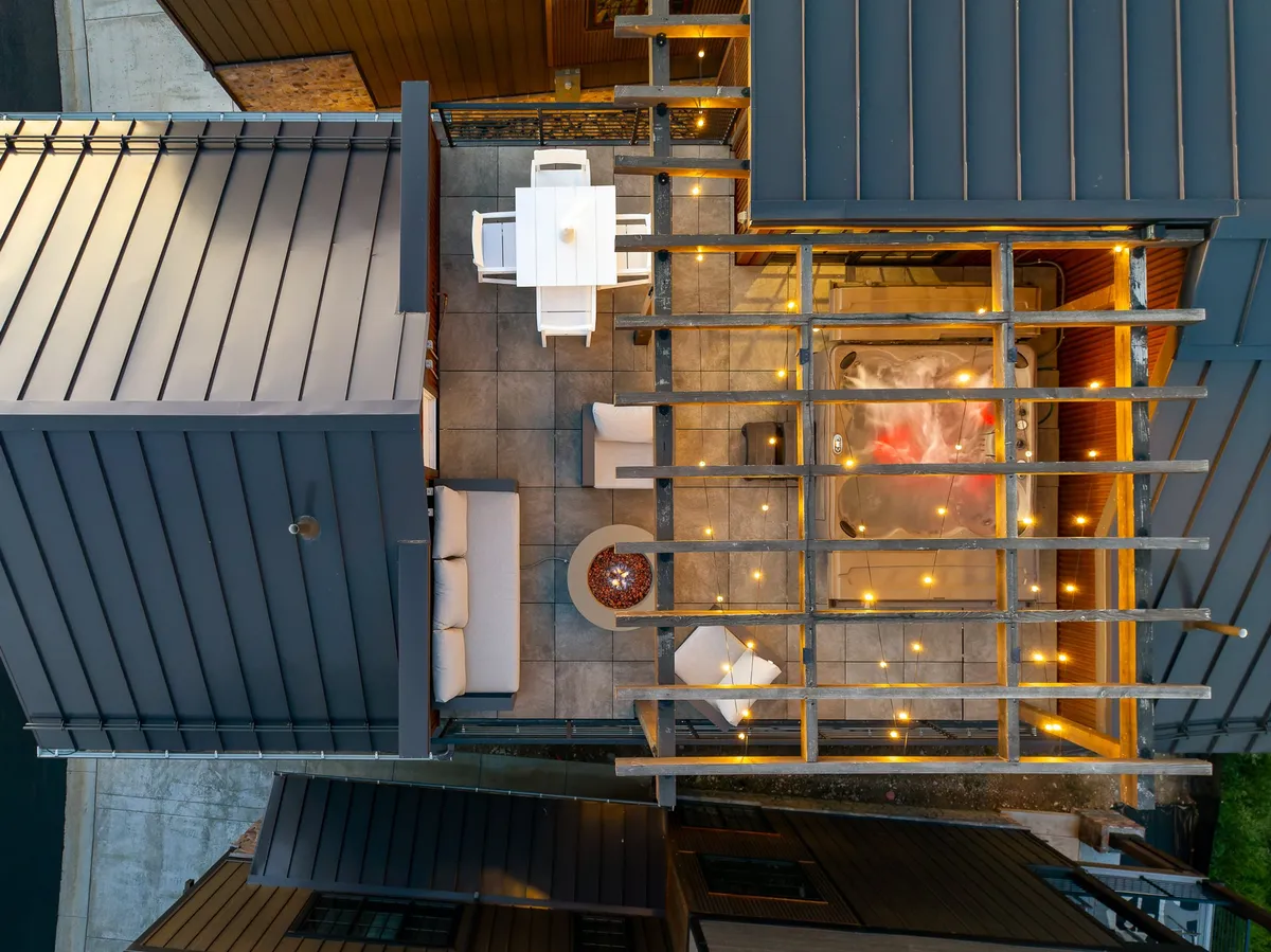 An inviting exterior scene showcasing a hot tub, an outdoor dining table, cozy seating areas, and string lights overhead. The setting exudes warmth and a welcoming ambiance for evening relaxation or gatherings.