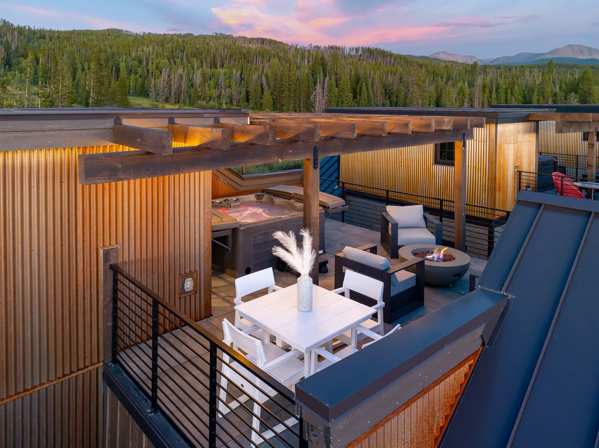 A spacious rooftop outdoor area featuring a dining table, a bubbling hot tub, a cozy seating area with a glowing fire pit, and sweeping panoramic mountain views. The sun is setting, casting warm light over the scene and inviting guests to relax, socialize, or soak in the luxurious ambiance.