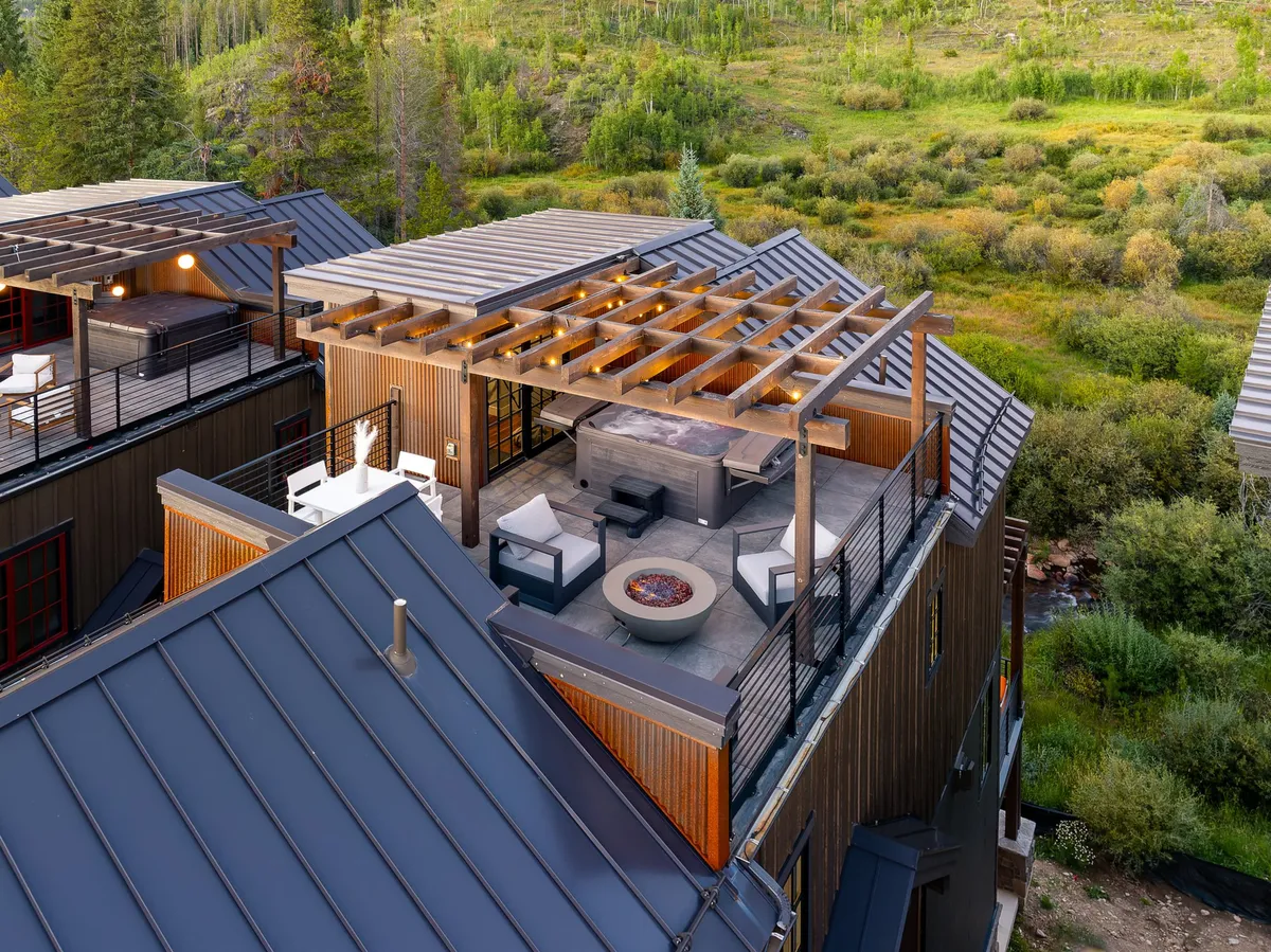 A spacious outdoor terrace featuring a private hot tub, a cozy fire pit, comfortable seating, and a pergola structure. The area is designed for relaxation and social gatherings, with mountain views in the background.