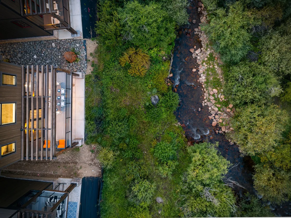 A modern deck overlooking a gently flowing stream and lush greenery, with an outdoor dining area offering a tranquil spot to connect with nature. The natural setting suggests easy access to riverside experiences and scenic adventure.