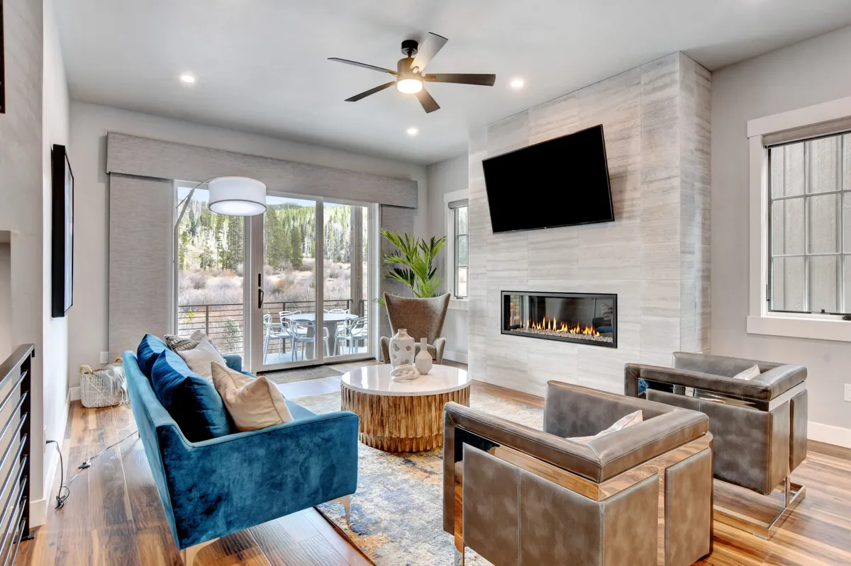 A spacious, sunlit living room featuring a plush blue sofa, modern coffee table, grand fireplace, and large windows that flood the space with natural light. The TV is positioned for easy viewing, and the overall layout is open and inviting, perfect for hosting gatherings or cozy nights in.