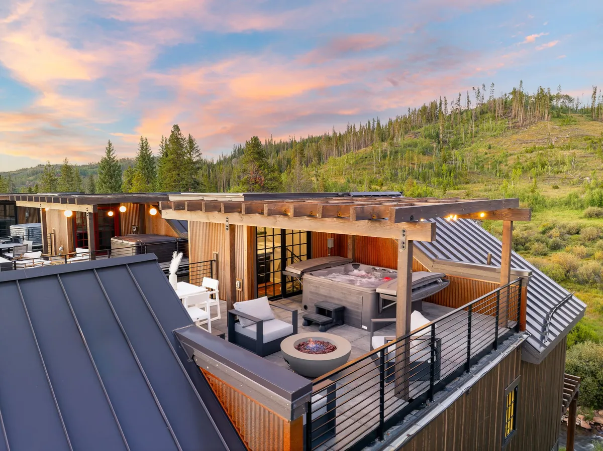 The image features the exterior of a luxury riverfront villa nestled amidst the scenic mountains of Winter Park, Colorado. The focal points are the spacious rooftop deck with a bubbling hot tub, a cozy fire pit surrounded by plush outdoor furniture, and panoramic views of the natural landscape. The setting is perfect for relaxing, socializing, watching sunsets, and stargazing, with ample space for groups to dine and unwind together.
