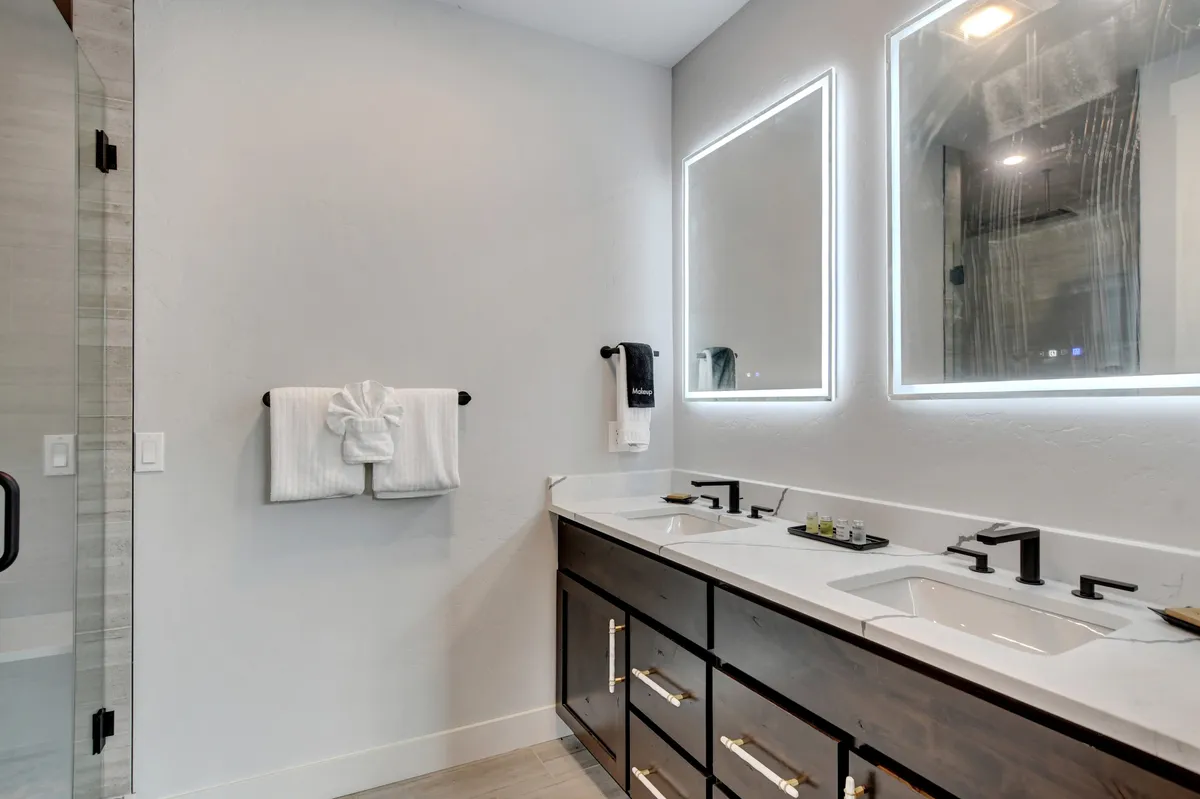 An elegant bathroom featuring dual sinks, illuminated mirrors, a shower area, and dark cabinetry. The space feels tranquil and sophisticated, ideal for self-care and relaxation.